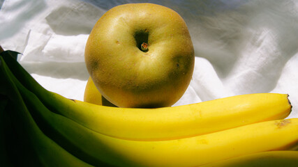 Ripe banana and green renet apples with aromatic laurel herbs, gifts of nature on white linen fabric, fruits for snacking, soft sunlight, seasonal fruits