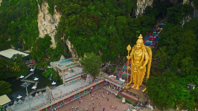 Scenic aerial footage of Batu Caves in Malaysia with the golden Lord Murugan statue, colorful temple complex, and rainbow stairs set against dramatic limestone cliffs.
