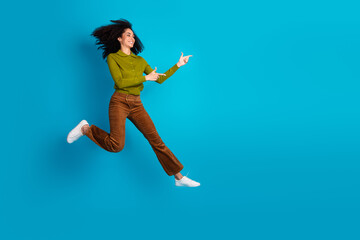 Fototapeta premium Energetic young woman in fashionable outfit jumping against vibrant blue background with charming smile