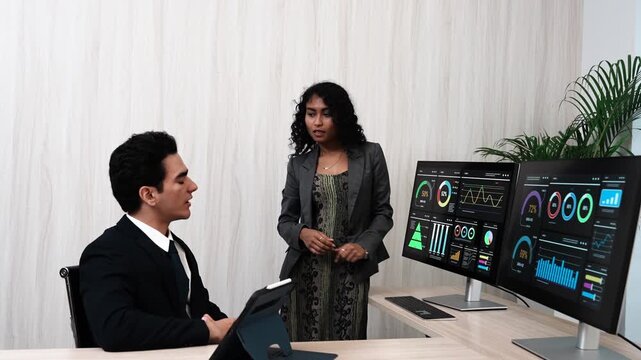 Businessman in suit sits at monitor screen writing software code and Asian female coworker discusses safety in using computer application at office desk.
