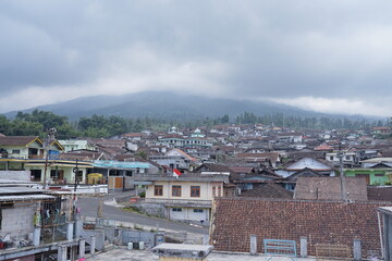 Obraz premium Sunday, ‎03 ‎August ‎2025, ‏‎14.20.00, Temanggung, Indonesia _ A scenic panorama of a traditional Asian village with terracotta roofs nestled at the base of a majestic mountain shrouded in mist