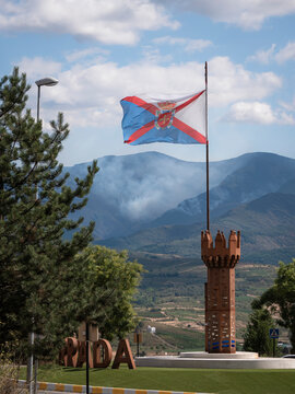 A flag of El Bierzo flies from a flagpole on a decorative tower while a mountainside burns in the background, causing large curtains of smoke during the devastating wildfires of 2025