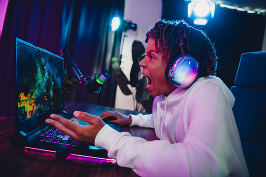 Teen gamer in headset streaming video games at night in neon-colored home studio setup with computer and microphone - Powered by Adobe