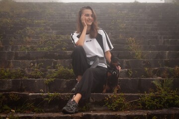 Fototapeta premium Young woman sitting on old stone steps in a foggy mountain landscape, wearing casual clothes and smiling softly. Atmospheric travel lifestyle photo.