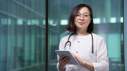 Portrait of smiling asian female doctor in white coat standing at workplace in modern hospital clinic office. Happy woman medical worker physician in glasses looking at camera holding tablet in hands - Powered by Adobe