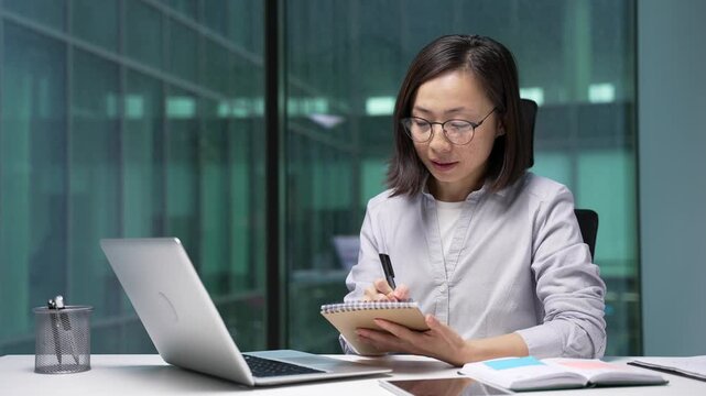 Asian businesswoman watching video call online conference taking notes in notebook looking at laptop screen sitting at workplace in office. Focused female employee listening remote business training