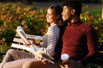 Happy multi-ethnic couple relaxing on bench reading books and drinking coffee in autumn park