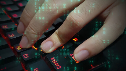 Close-up of a hand typing on a backlit gaming keyboard with red lights. An overlay of green binary code represents hacking, programming, and cybersecurity in the digital world of technology.