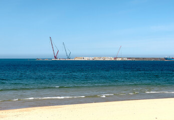 Beach with sand and blue sea waves. Horizon with cranes and industrial construction on pier by ocean
