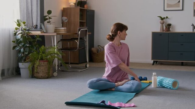 Full shot of elderly Caucasian woman with red hair, in pink t-shirt and blue leggings sitting on floor on fitness mat at home in half lotus posture, warming up back muscles with spine twist movements