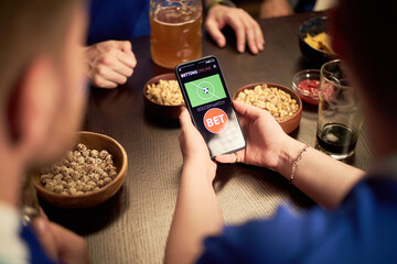 Group of young anonymous men watching soccer match together, one holding smartphone with online betting app, snacks and drinks on table, engaging in sports fan activity