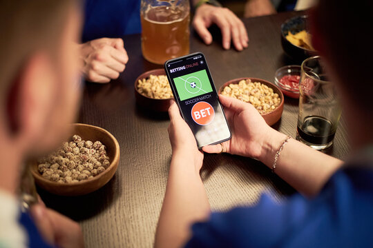 Group of young anonymous men watching soccer match together, one holding smartphone with online betting app, snacks and drinks on table, engaging in sports fan activity