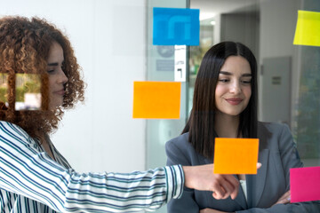 Businesswomen planning strategy using sticky notes on glass wall in office