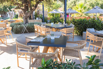 A stylish outdoor dining space featuring modern woven chairs with gray cushions, set tables with plates, glasses, and small potted plants, all arranged under a sleek pergola. 