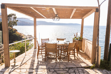 An inviting outdoor restaurant terrace with a wooden pergola and woven chairs, set against the backdrop of the sparkling blue Ionian Sea under the warm evening sun.
