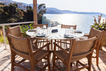 Scenic outdoor dining table on a sunlit terrace with panoramic sea views. Woven chairs and minimalist tableware create a relaxed yet refined atmosphere, ideal for vacation, hospitality.