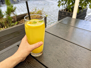 A hand holds a tall glass of refreshing orange drink with lemon slice and mint on a wooden outdoor table