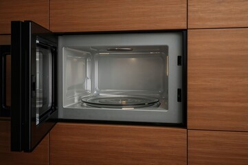 Fototapeta premium A sleek black microwave with its door open, built into a wooden kitchen cupboard. Inside, an empty glass turntable for heating food, shown from the side.