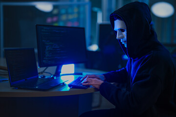 Obraz na płótnie Canvas A mysterious hooded hacker working on a computer in a dark room, illuminated by blue screen light. A concept for cybersecurity, hacking, cybercrime, and data privacy threats.