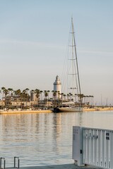 Malaga port with luxury yachts docked, the commercial area Muelle Uno, and the iconic lighthouse La Farola under a clear Mediterranean sky