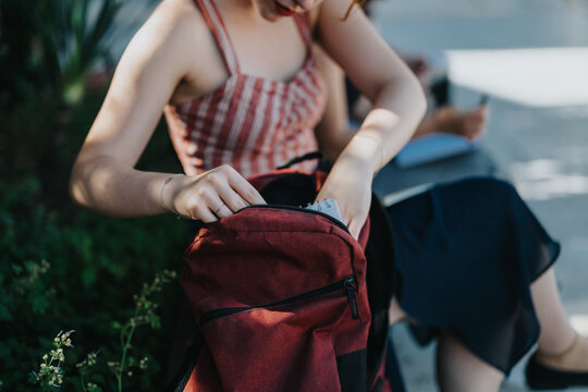 A young individual opening their red bag while seated on a bench outdoors on a bright day. The setting suggests a relaxed and natural environment, possibly in a courtyard or open area.