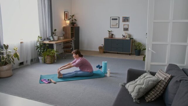 Wide shot of elderly 60-year-old retired Caucasian woman in pink t-shirt and blue leggings doing forward bend, then standing up into trikonasana posture, while exercising alone in living room at home