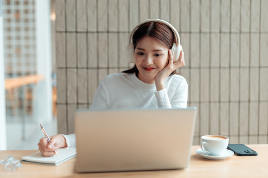 Asian freelancer woman taking notes while attending a video conference from home using laptop and headphones
