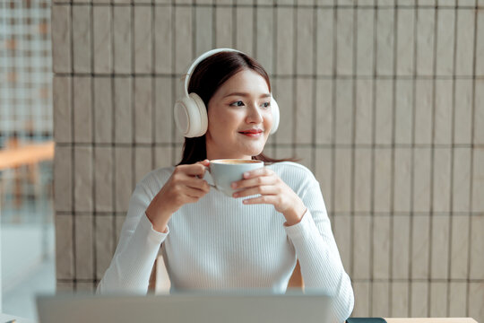 Happy businesswoman wearing headphones and drinking coffee while working on laptop computer in cafe - Powered by Adobe