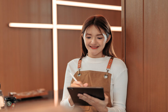 Asian female waitress wearing apron using tablet and stylus pen taking orders from customer in coffee shop