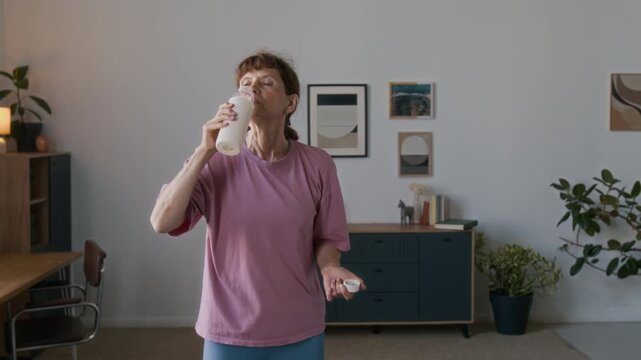 Medium arc shot of energetic sporty senior Caucasian woman in pink t-shirt standing in living room at home, drinking healthy protein shake or smoothie after home workout, while keeping fit in old age