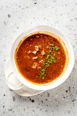 Traditional beef soup served in a white bowl with sour cream and herbs on a rustic stone background. Classic Eastern European comfort food presentation