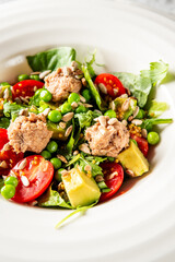 Fresh salad with cherry tomatoes, avocado, spinach, peas, and tuna on a white plate over gray countertop. Healthy meal concept, top view
