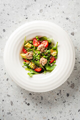 Fresh salad with cherry tomatoes, avocado, spinach, peas, and tuna on a white plate over gray countertop. Healthy meal concept, top view