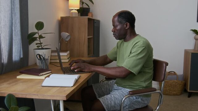 Medium shot of mature African American man sitting at work desk at home, typing on laptop, stretching, massaging stiff neck, bending and rotating head, then standing up to take break