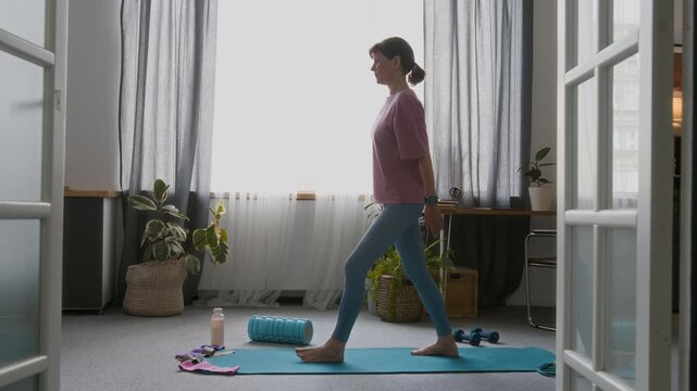 Full shot of slim beautiful senior Caucasian woman in pink t-shirt and blue leggings practicing smooth tai hi routine in living room at home, while standing against bright light from window