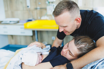 A man hugs his wife and newborn baby in the delivery room. The woman gave birth and the man is...