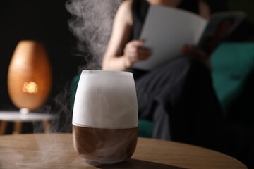 Woman reading magazine in armchair indoors, focus on modern aroma lamp