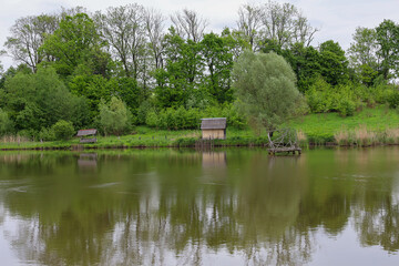 Fototapeta premium A serene landscape with green trees and grass reflecting in calm water. Wooden fishing huts are located near the shore, creating an idyllic atmosphere.