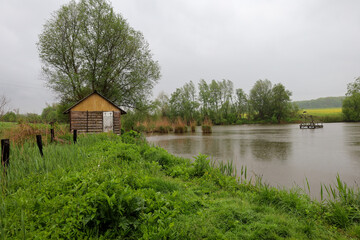 Fototapeta premium ​A cozy wooden house is located on the bank of a pond under a gray, rainy sky. The surrounding greenery and the reflection of trees in the water create an atmosphere of peace and detachment from the d