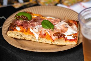 A slice of pizza topped with fresh basil leaves, prosciutto crudo and melting stracciatella. The pizza is served on a paper plate with a glass of beer