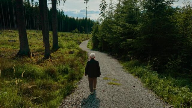 Pensioner walking down forest path enjoying quiet scenery. Older woman strolling through woodland trail feeling calm. Senior woman walking along gravel road embracing nature