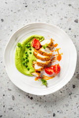 Grilled chicken breast with orange sauce, green pea puree, cherry tomatoes, and parsley on a white plate. Elegant gourmet presentation on a stone surface