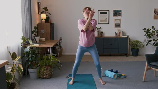 Full shot of senior Caucasian woman in pink t-shirt and blue leggings standing on yoga mat, warming up with arm and leg movements to practice qigong or tai chi routine at home