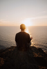 young man meditating on rocky cliff at sunset overlooking ocean for tranquility and mindfulness