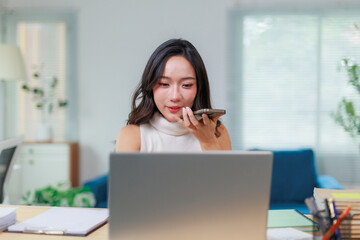 Businesswoman recording a voice message on her smartphone while working diligently on her laptop in a cozy home office setting