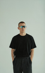 Fototapeta premium A minimalist studio portrait against a plain light gray background. The subject wears a plain black crew neck t-shirt and dark gray loose-fitting pants.