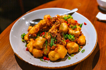 Chinese-style deep-fried tofu topped with chopped green onions, red chili pieces, and fried garlic, served in a white ceramic bowl on a wooden table