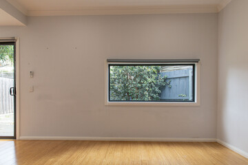 Minimalist room with light wooden flooring, large horizontal window with a view of a leafy tree and black fence, and soft natural light entering from outside