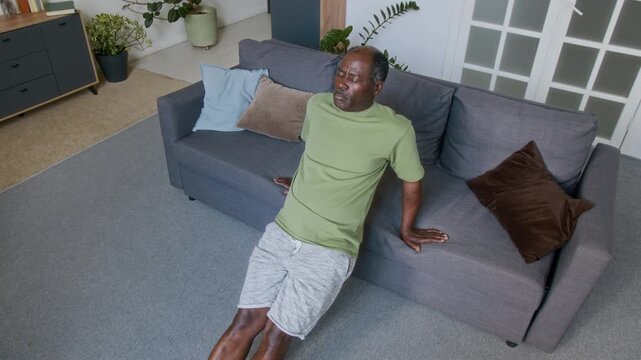 Medium high-angle shot of determined senior African American man performing repeats of tricep press exercise, leaning on seat of couch in living room, while keeping fit during retirement