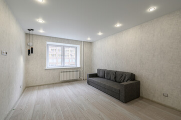 bright, empty room with a grey sofa. There's a window with a radiator below. The walls are textured, the floor is light wood. Spotlights illuminate the space